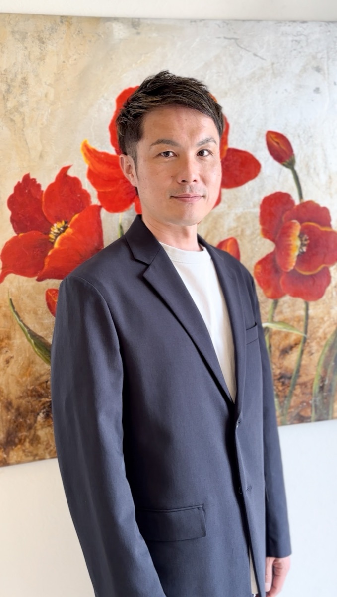 ジャケット姿で正面を向く原口卓也の写真 背景は赤い花の絵